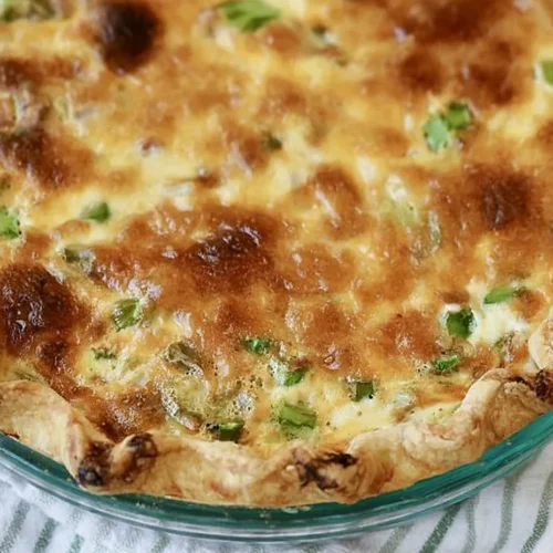 Close-up view of a baked asparagus and bacon quiche in a glass pie pan, highlighting the golden-brown blistered cheese surface, visible green asparagus pieces, and flaky pastry crust.