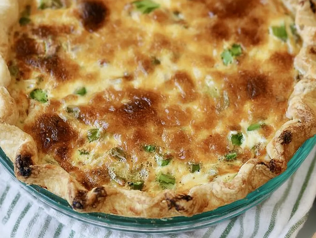 Close-up view of a baked asparagus and bacon quiche in a glass pie pan, highlighting the golden-brown blistered cheese surface, visible green asparagus pieces, and flaky pastry crust.