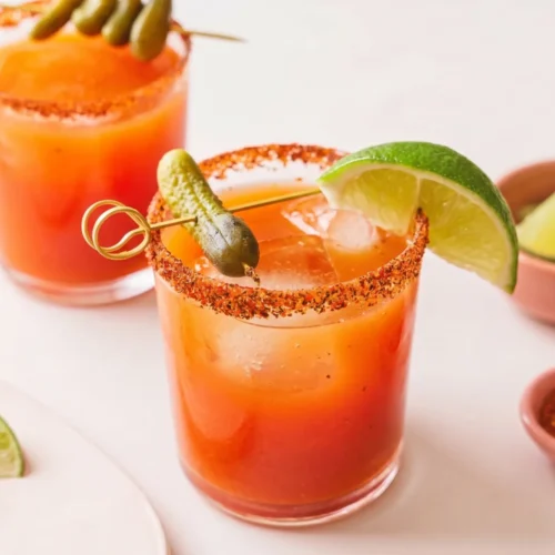 Two glasses of bright orange-red michelada mocktails served over large ice cubes on a light surface. The rims of the glasses are heavily coated in red Tajín seasoning. The front drink is garnished with a fresh lime wedge and a gold cocktail pick holding a small green cornichon pickle. Small, rustic bowls containing lime wedges and extra red spice sit softly out of focus in the background.