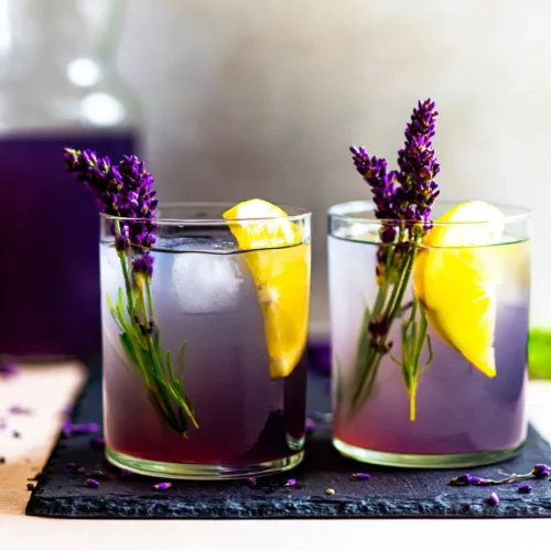 Two short glasses of layered lavender lemonade mocktails on a dark slate board, featuring a deep purple bottom layer and a clear top layer, garnished with fresh lemon wedges and tall purple lavender sprigs. A glass pitcher of the purple beverage rests in the blurred background.