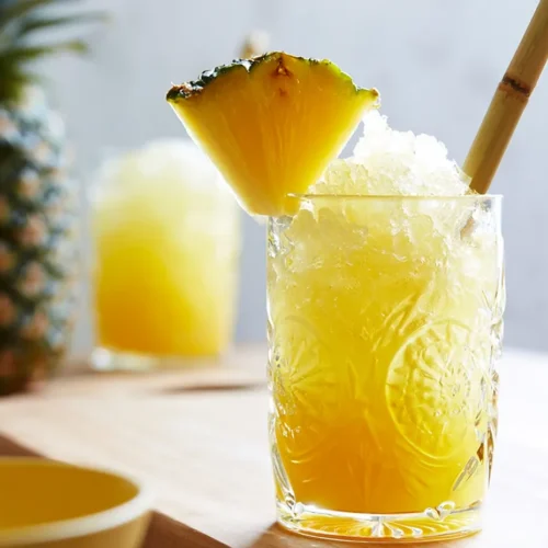 A close-up of an ornate vintage glass filled with bright yellow pineapple agua fresca and crushed ice, garnished with a fresh pineapple wedge and a bamboo straw. A whole pineapple rests in the blurred background.