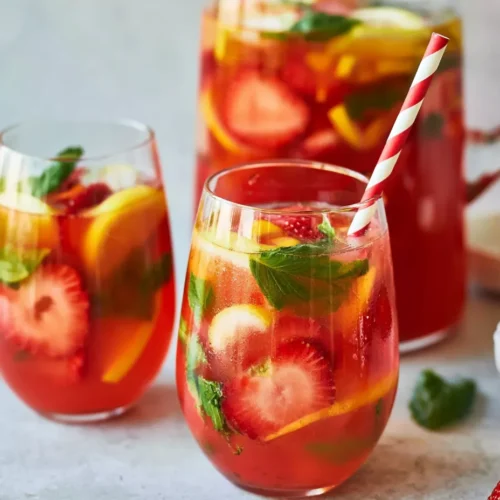 Two stemless glasses and a large pitcher filled with a vibrant red iced beverage, garnished with fresh sliced strawberries, lemon wedges, and green herbs, resting on a light gray surface.