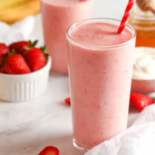 A close-up shot of a tall glass filled with a pale pink strawberry banana smoothie, featuring a red and white striped straw. The glass sits on a bright marble countertop surrounded by a white ramekin of fresh strawberries, a jar of honey with a wooden dipper, a small bowl of creamy yogurt, and a casually draped white linen napkin.