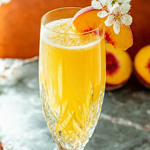 A close-up editorial shot of a vibrant, golden Peach Bellini mocktail served in a textured crystal flute. The sparkling drink is garnished with a fresh peach slice and delicate white blossoms, surrounded by halved fresh peaches on a dark marble surface.