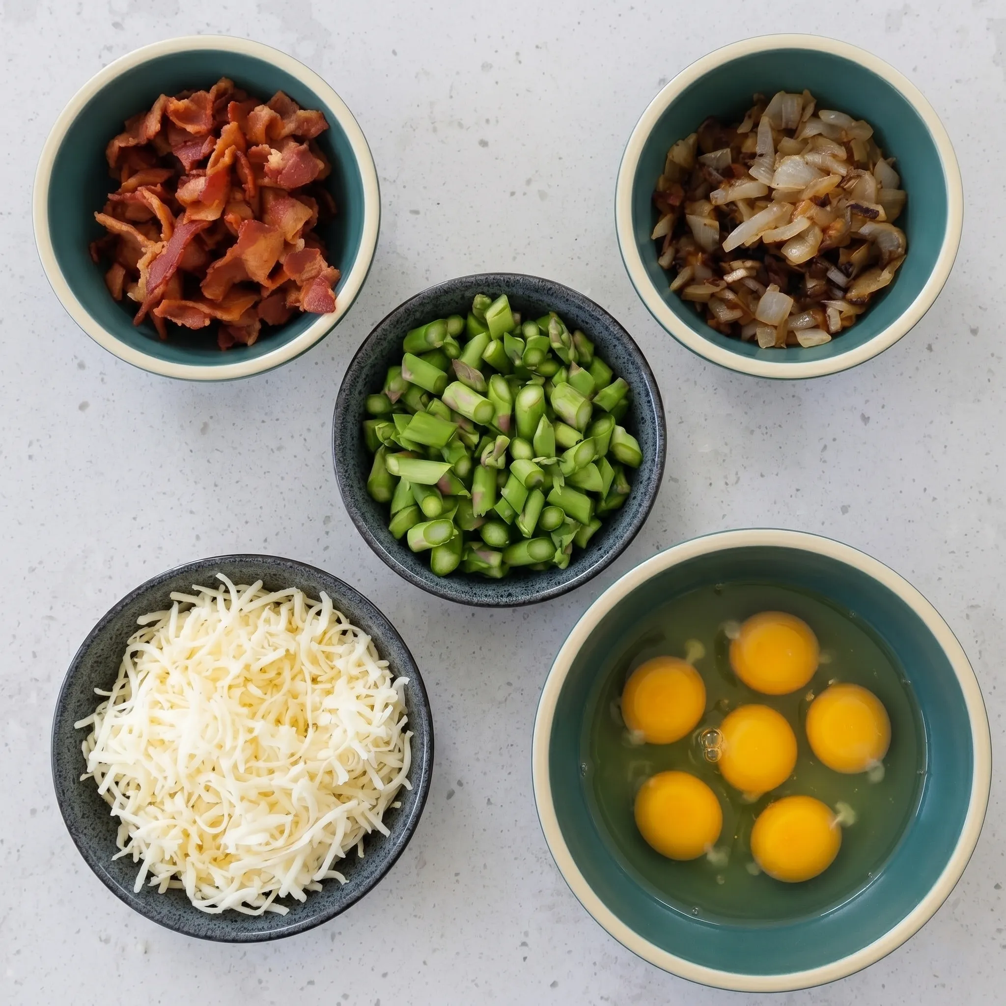 Prepped ingredients for asparagus and bacon quiche arranged in individual bowls on a light speckled countertop, including crumbled bacon, sautéed onions, chopped green asparagus, shredded Gruyère cheese, and cracked raw eggs.