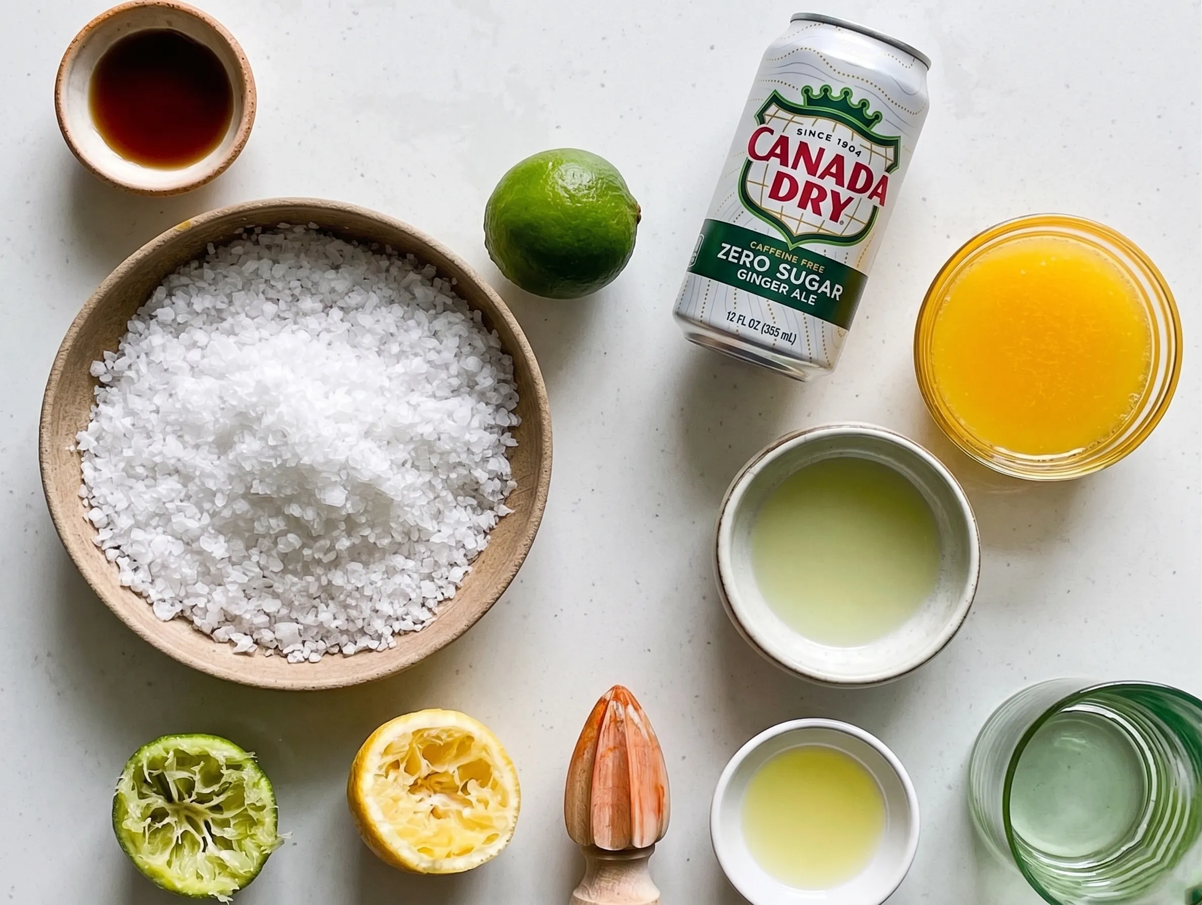 An overhead view of ingredients for a virgin margarita mocktail arranged on a light countertop, including a bowl of coarse sea salt, a whole lime, squeezed lime and lemon halves, small bowls of fresh citrus juices and agave nectar, a wooden citrus reamer, and a can of sparkling zero-sugar ginger ale.