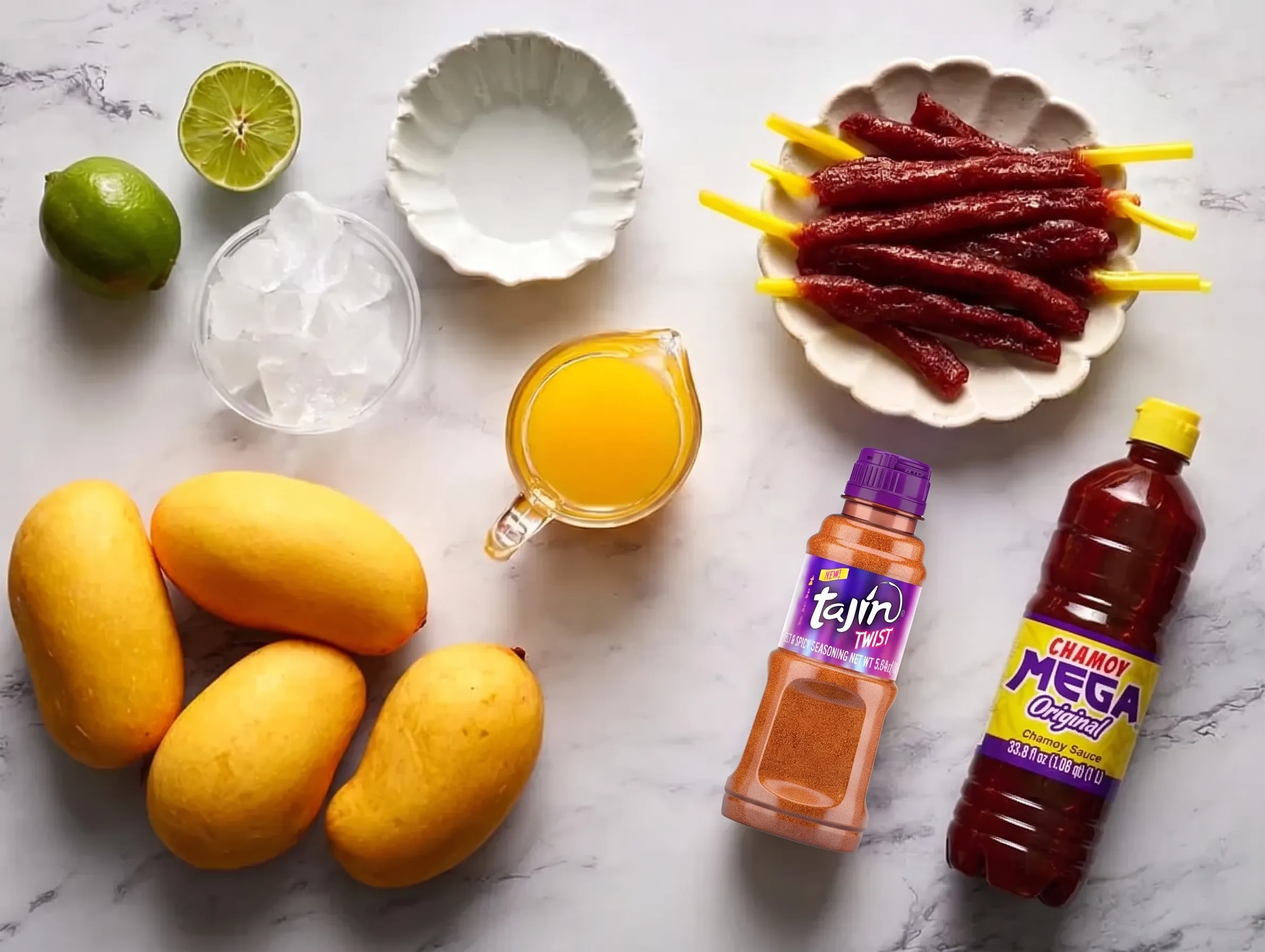 An overhead shot of mango chamoy mocktail ingredients arranged on a white marble surface, featuring four fresh yellow mangoes, whole and halved limes, a bowl of ice, a small pitcher of mango juice, a bottle of Tajín Twist seasoning, a bottle of Mega Original Chamoy Sauce, and a plate of tamarind candy straws.