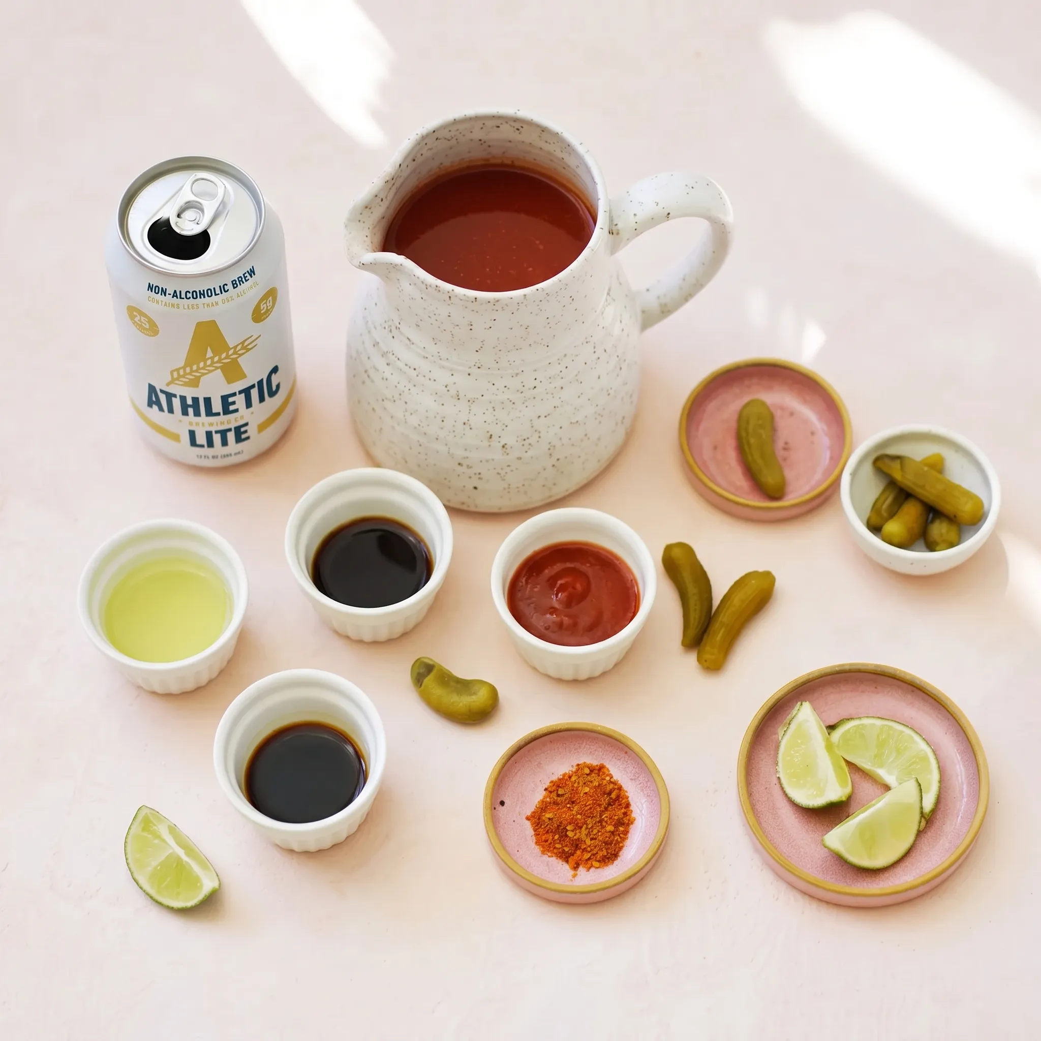 A flat lay of michelada mocktail ingredients arranged on a light pink surface. The collection includes a can of Athletic Lite non-alcoholic beer, a rustic speckled pitcher filled with vibrant red tomato juice, and several small bowls containing fresh lime juice, dark tamari, Worcestershire sauce, red hot sauce, Tajín seasoning, cut lime wedges, and small green cornichon pickles.