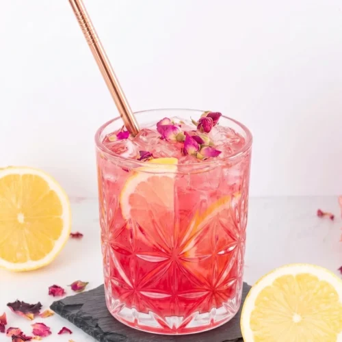 A close-up of a bright pink rose lemonade mocktail in a clear, textured glass filled with ice, lemon slices, and dried rose buds. A sleek copper straw rests in the drink, which sits on a square dark slate coaster surrounded by fresh lemon halves and scattered dried rose petals against a bright white background.