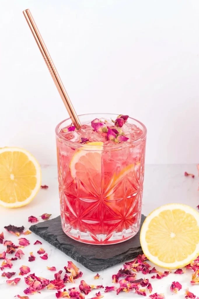 A close-up of a bright pink rose lemonade mocktail in a clear, textured glass filled with ice, lemon slices, and dried rose buds. A sleek copper straw rests in the drink, which sits on a square dark slate coaster surrounded by fresh lemon halves and scattered dried rose petals against a bright white background.