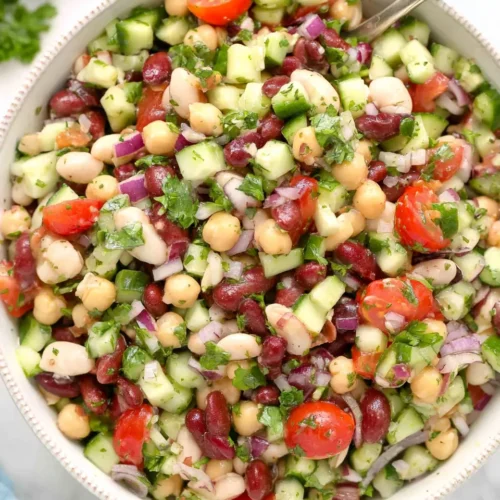 Close-up overhead view of a large white bowl filled with Mediterranean Bean Salad, containing a mix of chickpeas, kidney beans, white beans, diced cucumbers, cherry tomatoes, red onions, and fresh parsley, with a metal serving spoon resting inside.