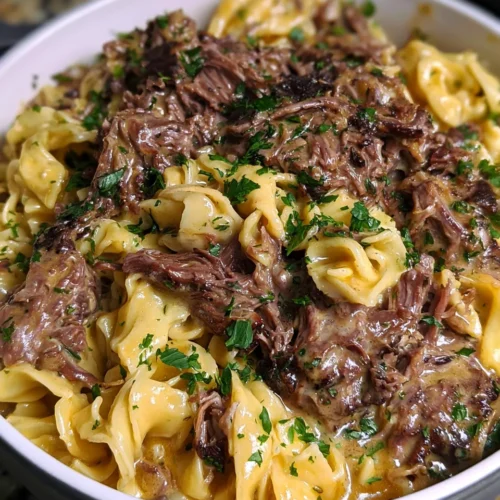 Close-up of a white bowl filled with Crock Pot Beef and Noodles, featuring tender shredded beef and gravy served over wide egg noodles, garnished with chopped fresh parsley.
