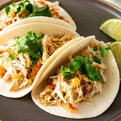 Close-up view of three shredded chicken tacos in soft flour tortillas on a dark gray plate, garnished with shredded white cheese and fresh cilantro, with two lime wedges resting on the side.