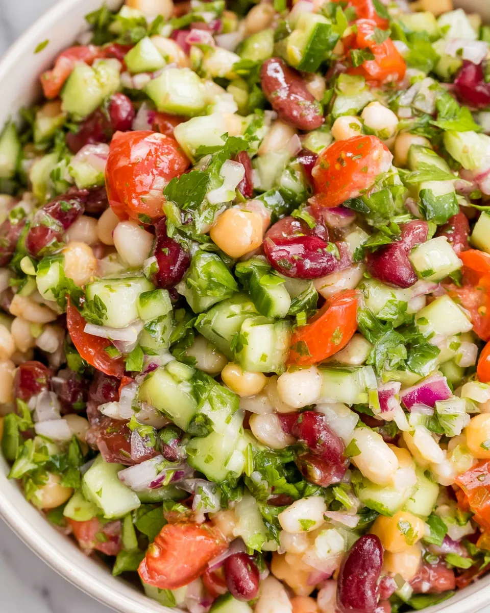 freshly tossed Mediterranean Bean Salad Extreme close-up of a colorful Mediterranean bean salad in a light-colored bowl, showing a glossy mixture of kidney beans, chickpeas, white beans, diced cucumbers, halved cherry tomatoes, red onions, and chopped parsley.