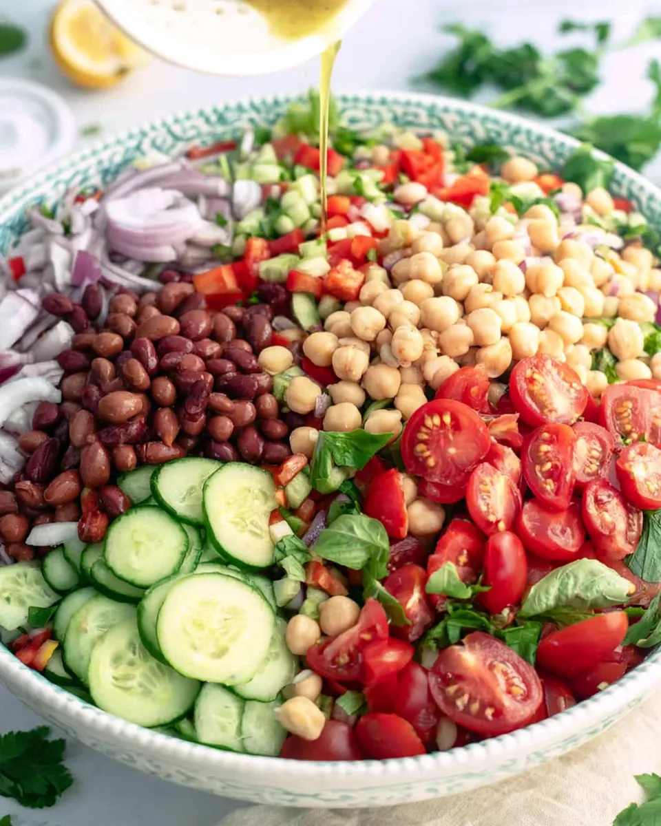 unmixed Mediterranean bean salad ingredients Close-up view of a large, patterned bowl filled with unmixed salad ingredients arranged in sections, including sliced red onions, black beans, chickpeas, halved cherry tomatoes, and sliced cucumbers, with a yellow vinaigrette dressing being poured over the top from a white bowl.