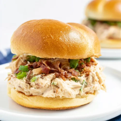 A close-up of an Instant Pot crack chicken sandwich on a toasted golden bun, filled with creamy shredded chicken, crumbled bacon, and green onions, sitting on a white plate over a blue and white checkered cloth.