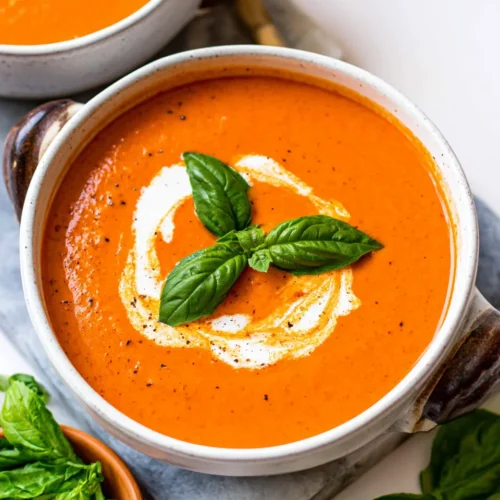 Close-up of a speckled ceramic bowl filled with vibrant orange Roasted Tomato Soup, garnished with a swirl of cream, a sprinkle of black pepper, and a sprig of fresh basil. The bowl sits on a grey stone board with fresh basil leaves nearby.