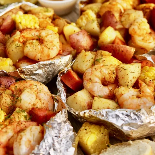 Several opened aluminum foil packets containing cooked shrimp, diced red potatoes, smoked sausage slices, and corn on the cob, all coated in a red spice blend and green herbs. Lemon wedges, a small bowl of dipping sauce, and crusty bread sit in the background.