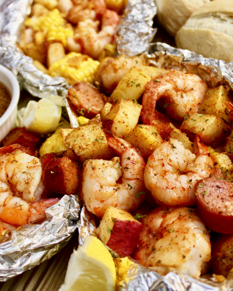 An open aluminum foil packet filled with seasoned, cooked pink shrimp, cubed red potatoes, sliced smoked sausage, and corn on the cob. Fresh lemon wedges and a small bowl of dipping sauce are visible in the background.