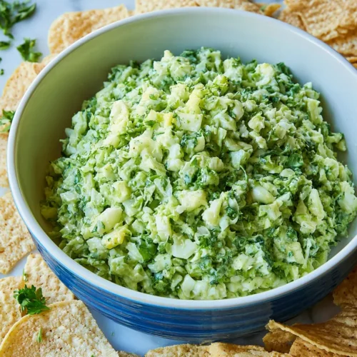 A blue bowl filled with a finely chopped, creamy green salad sits on a white marble surface, completely surrounded by a ring of tortilla chips and scattered fresh herbs.