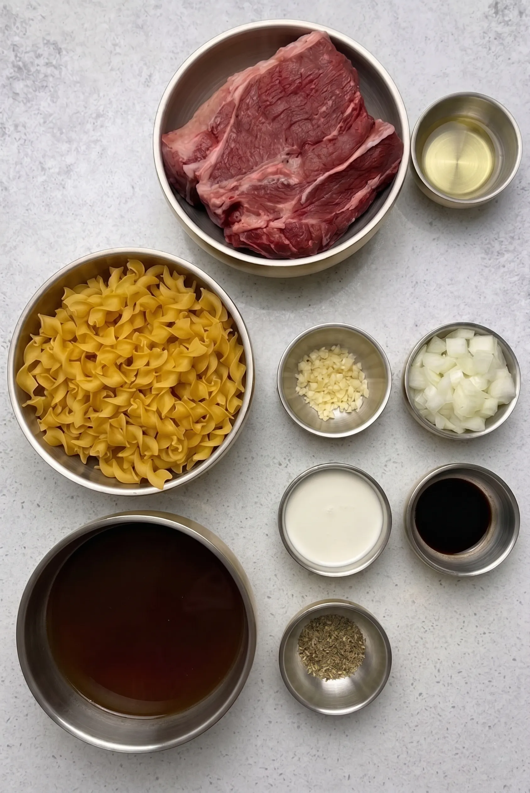 Overhead view of prepped ingredients for Crock Pot Beef and Noodles arranged in stainless steel bowls on a light speckled countertop. The bowls contain a raw beef roast, uncooked egg noodles, beef broth, diced onions, minced garlic, cooking oil, a creamy white liquid, a dark sauce, and dried herbs.