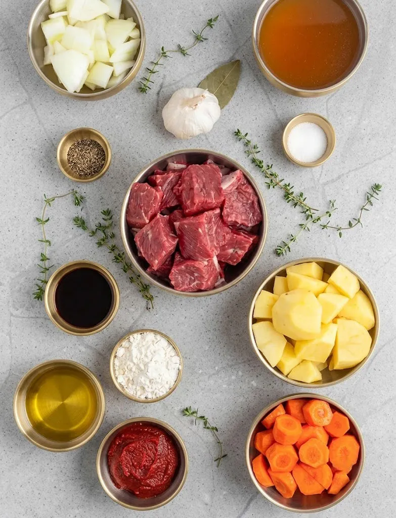 An overhead flat lay of raw beef stew ingredients neatly organized in small golden bowls on a gray textured countertop, including cubed beef, chopped onions, potatoes, carrots, broth, oil, tomato paste, flour, a dark liquid, salt, pepper, a whole head of garlic, a bay leaf, and sprigs of fresh thyme.