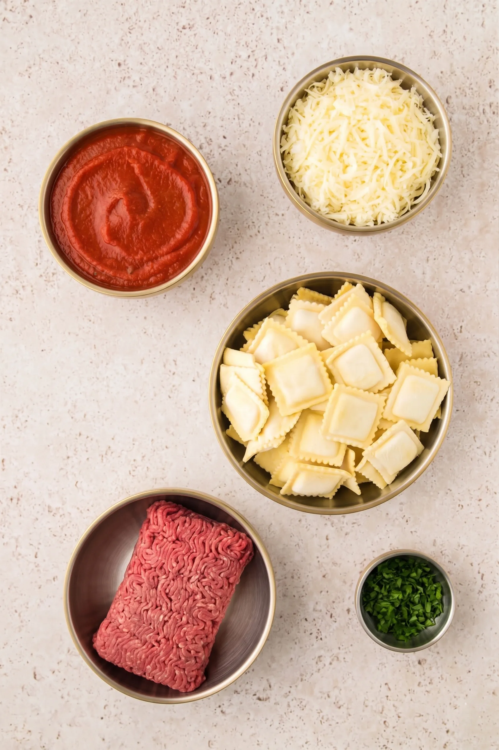 Ingredients for a ravioli dish: ground beef, cheese, ravioli, marinara sauce, and chopped herbs in bowls.