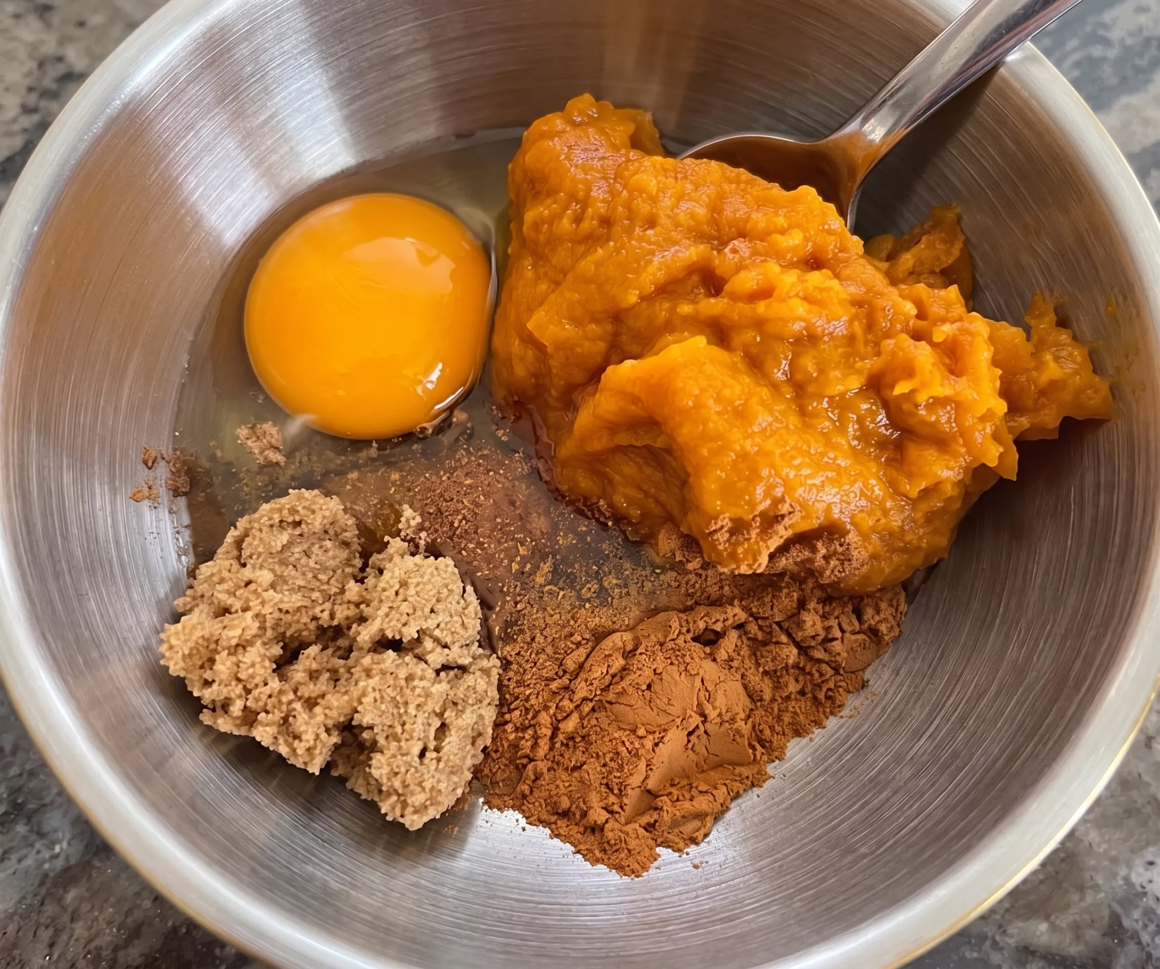 Pumpkin puree, an egg, brown sugar, and spices in a mixing bowl, ready for baking preparation.