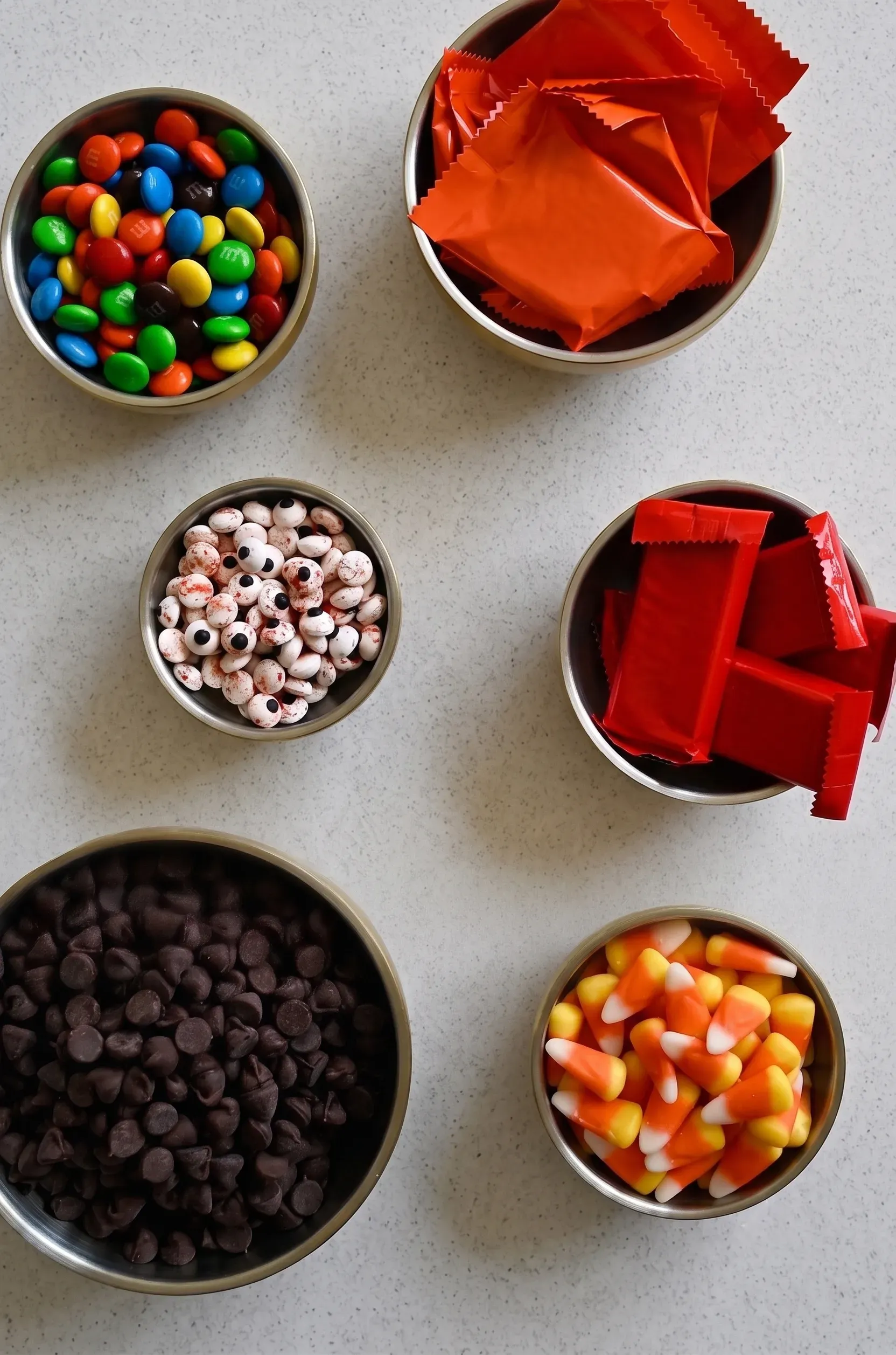 ingredients Colorful candy selection including M&Ms, candy corn, and chocolate chips in bowls on a light countertop.